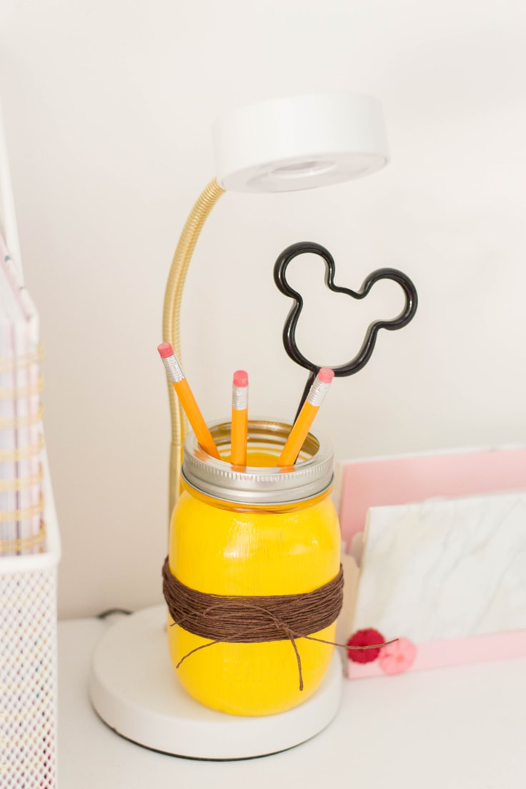 The DIY mason jar pencil holder sitting on a desk with a desk lamp over it. 