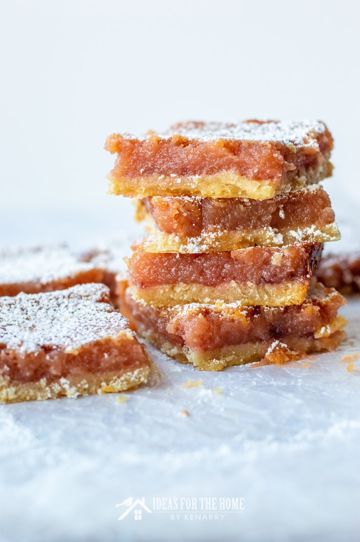 Side view of sliced raspberry lemon bars. 4 of these desserts are stacked on top of one another. There are a few single raspberry lemon bars near by. All of these delicious desserts are sprinkled with powdered sugar.