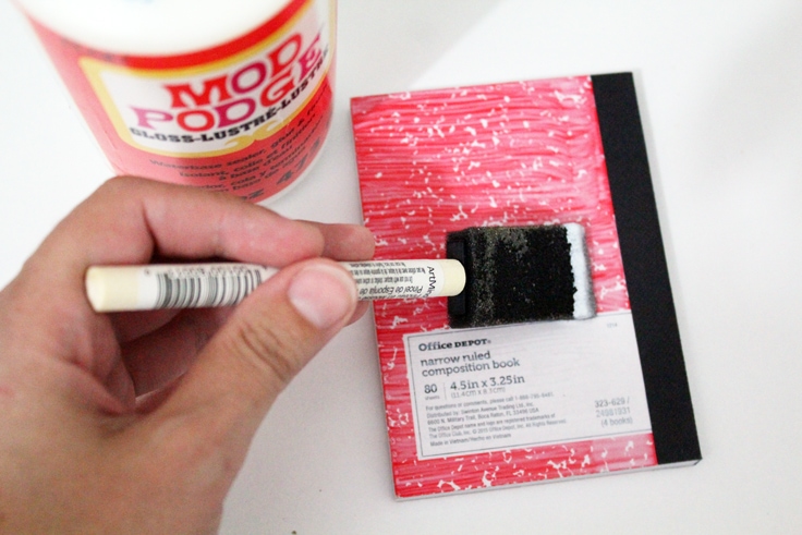 A hand using a sponge to brush Mod Podge onto the notebook