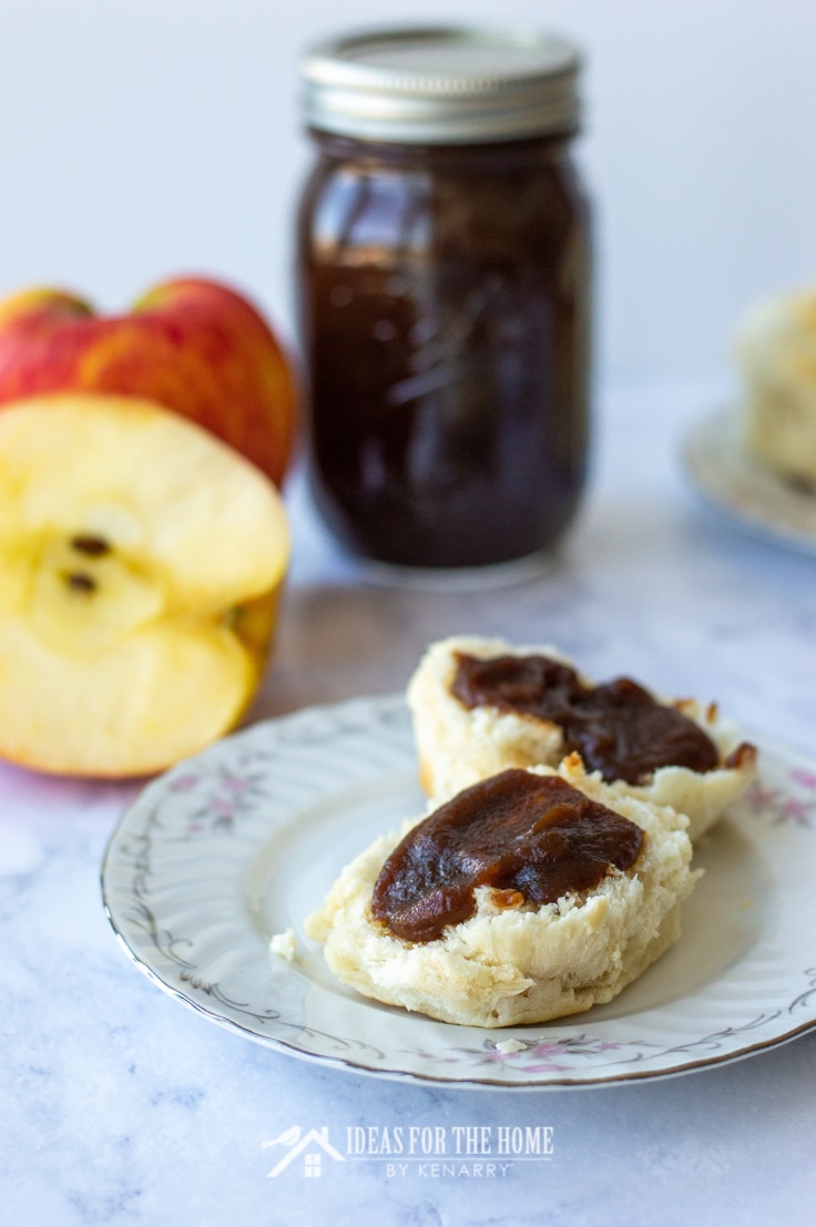 A warm homemade biscuit on a plate. The biscuit is split in half with delicious apple butter smothered on each side. There's a fresh apple and a full jar of the butter in the background.