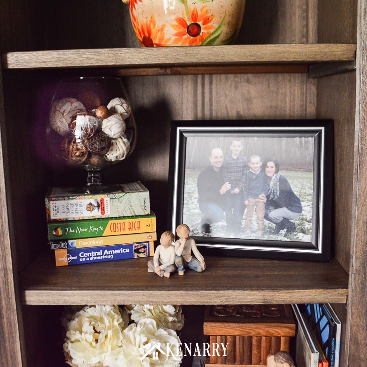 A glass vase filled with decorative balls on a stack of travel books next to a family photo and Willow Tree figurines of boys