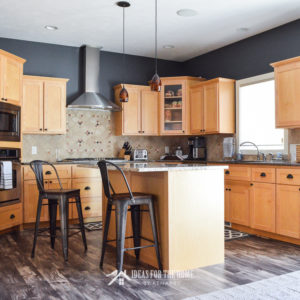 Kitchen reveal showcasing natural maple cabinets and Allen and Roth Chesterfield Hackberry laminate flooring. An industrial style metal bar stool with wood seat next to a kitchen island with Giallo Ornamental granite.