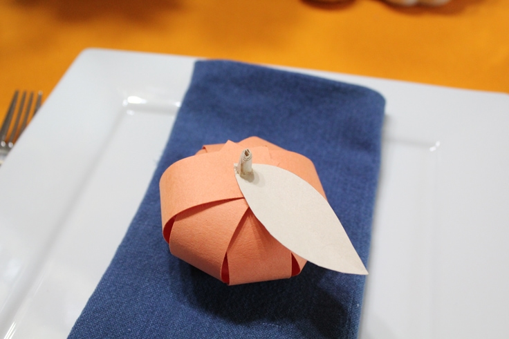 an orange paper pumpkin with a manila paper leaf and stem sitting on a blue napkin stacked on a white plate
