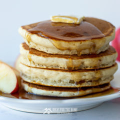 A stack of four cinnamon applesauce pancakes covered with butter and syrup along with sliced apples