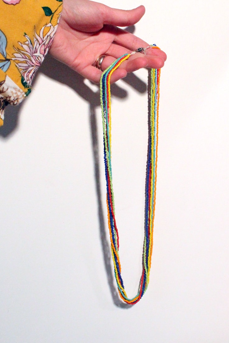 left hand holding a rainbow seed bead necklace in front of a white background