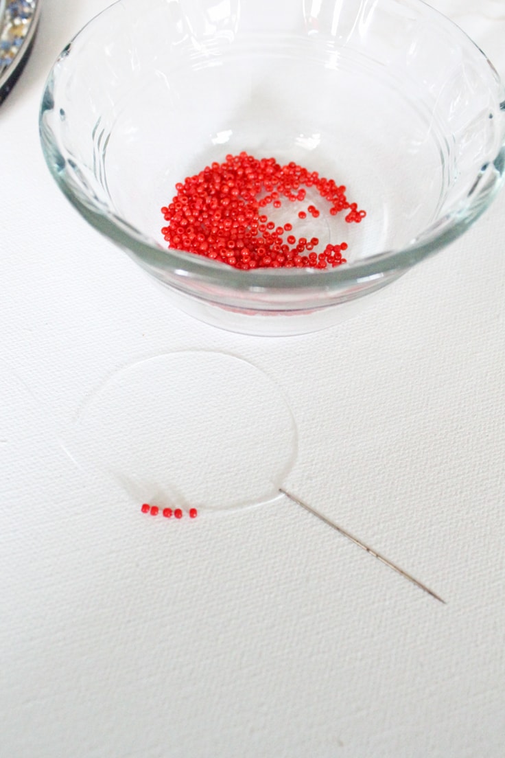glass bowl of red beads beside a threaded needle with more red beads