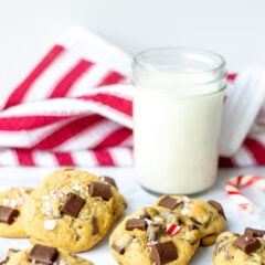 A glass of milk and candy canes with a pile of cookies topped with crushed candy and chocolate chunks