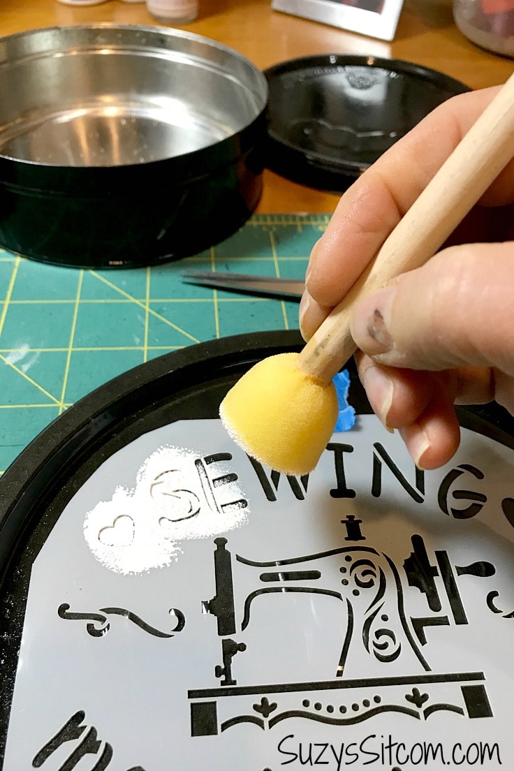 Painting the stencil on the cookie tin with white paint and a sponge brush