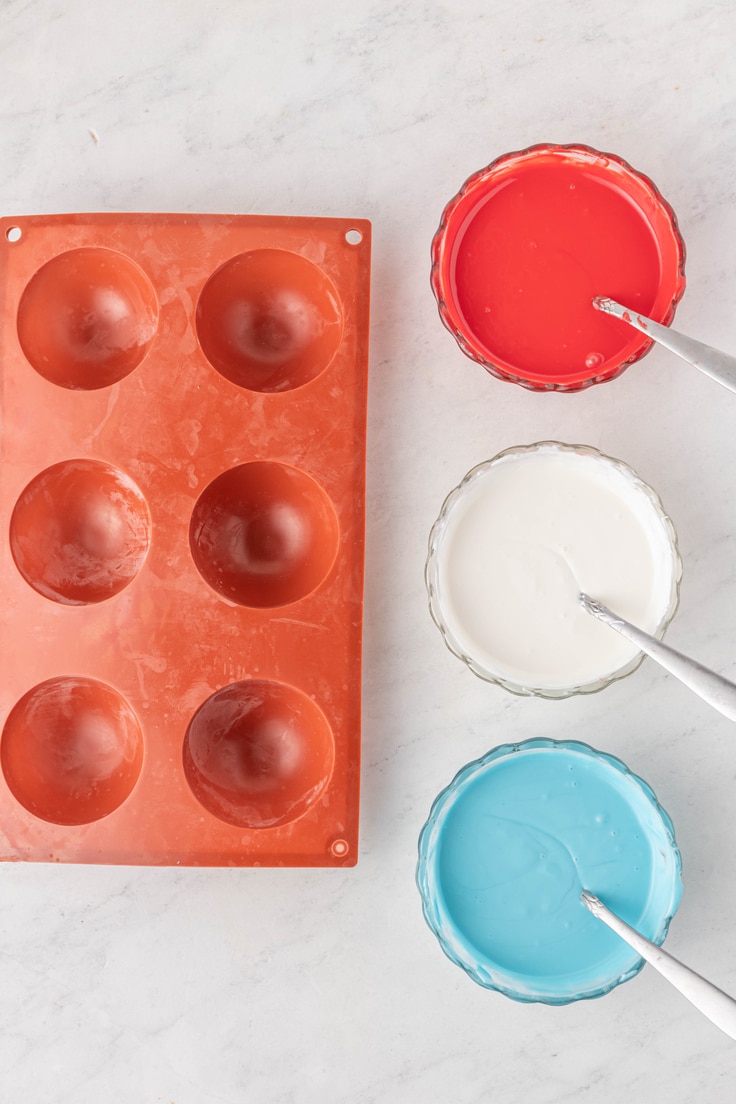 Sphere silicon mold beside bowls of melted chocolate in red, white, and blue.