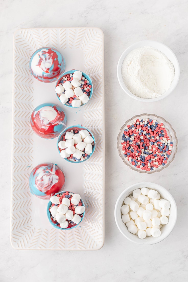 Half-opened hot cocoa bombs beside prep bowls full of white cocoa mix, patriotic sprinkles, and marshmallows.