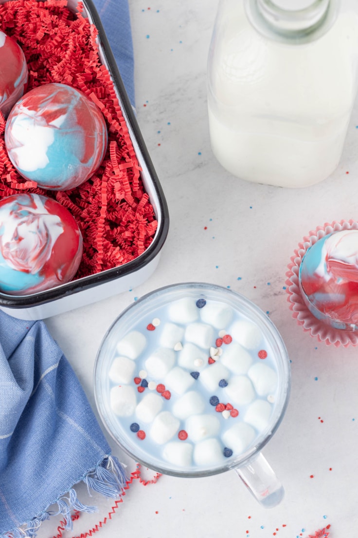 Red, white, and blue hot cocoa bombs beside a glass full of cocoa and marshmallows.