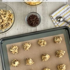 Drops of cookie dough on a pan with ingredients including potato chips and chocolate chips in the background.