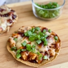 BBQ Chicken Tostada topped with green onion on a cutting board with a bowl of onions in the background.