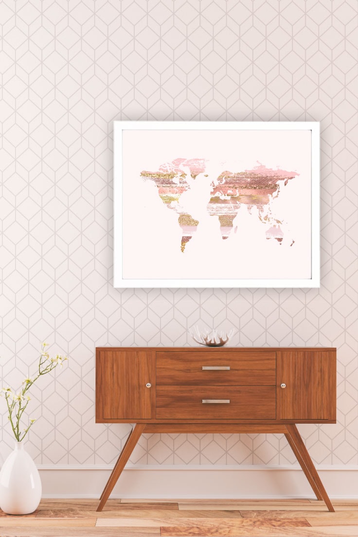 Pink glitter world map design in a white frame on geometric wallpaper above wooden drawer with white vase and ornamental flowers on the left.