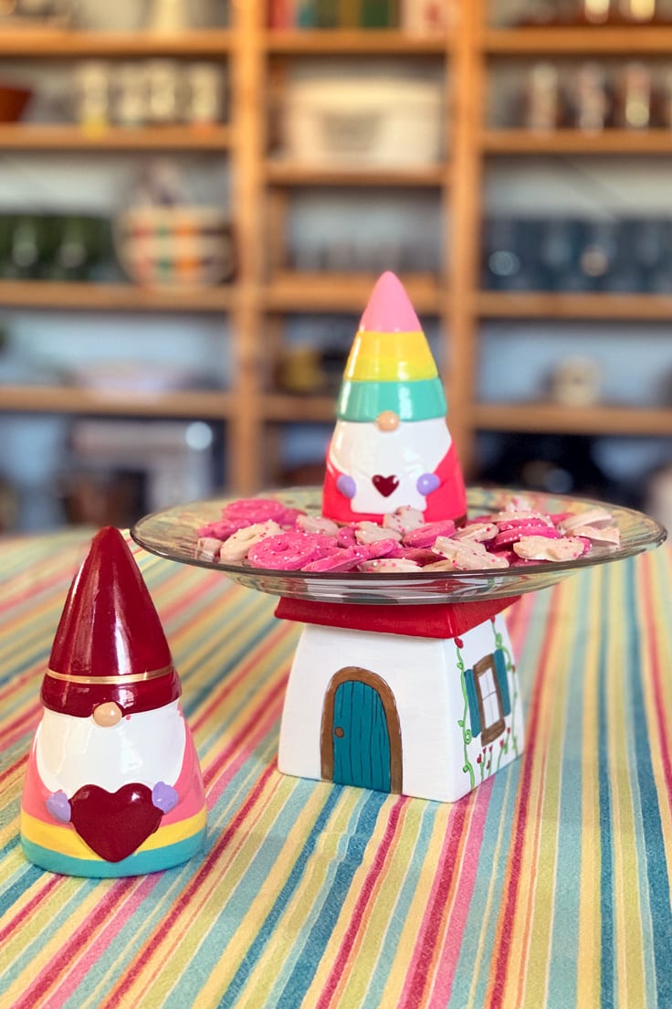 Two glass valentine gnomes standing beside a gnome house cake plate painted in red, blue, and gold.