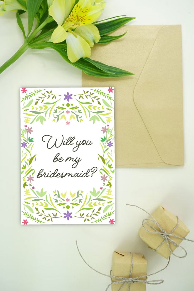Preview of bridesmaid card on white desk with kraft envelope and foilage on top and two kraft gift packages on the lower right.