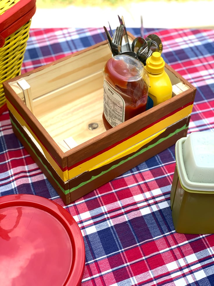 An outdoor cookout setup on a plaid tablecloth with plates, pickles, ketchup, and mustard.