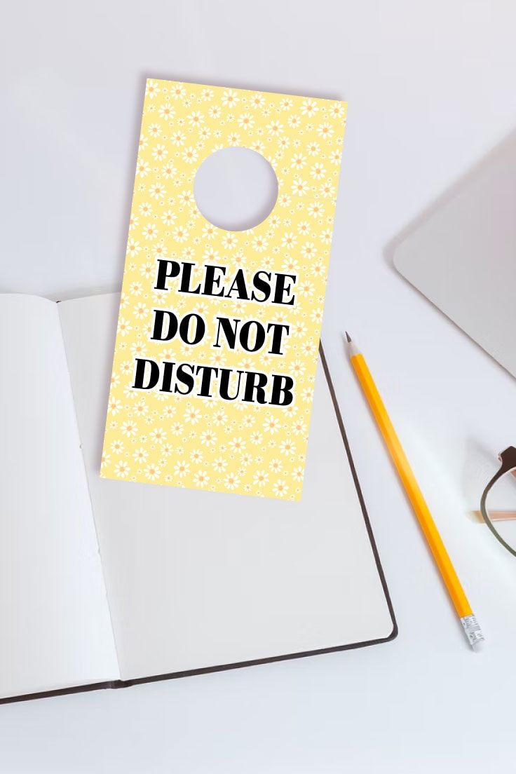Preview of yellow printable door hanger design on white desk with blank notebook, pencil and corner of laptop computer and glasses in view. 