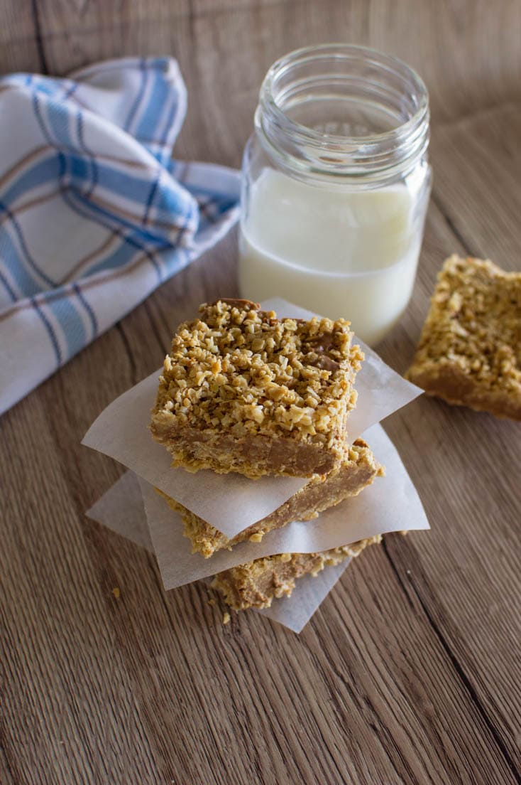 A stack of no-bake chocolate oatmeal bars with parchment paper in between, sitting on a wooden table, with a mason jar of milk in the background.