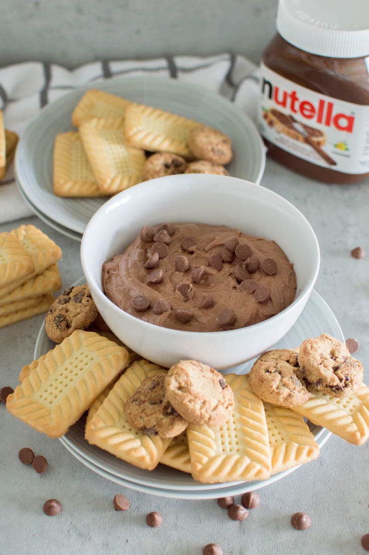 A bowl of Nutella dip, topped off with chocolate chips, surrounded by different kinds of cookies and a jar of Nutella in the background