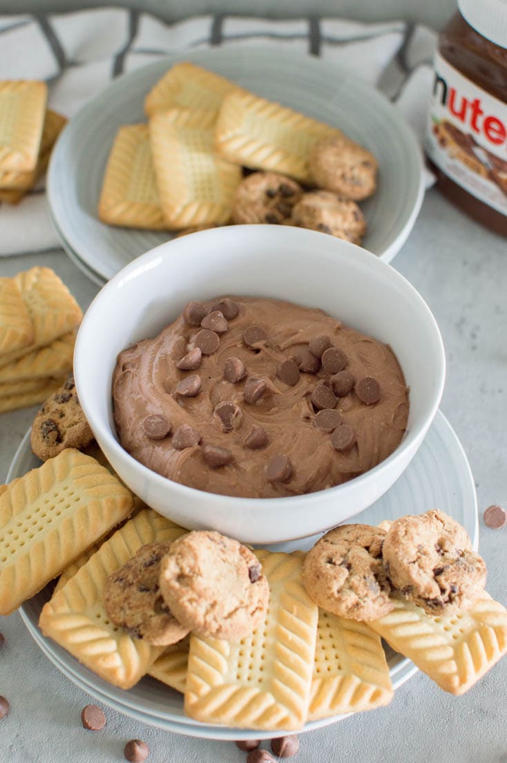 A bowl of Nutella dip, topped off with chocolate chips, surrounded by different kinds of cookies and a jar of Nutella in the background