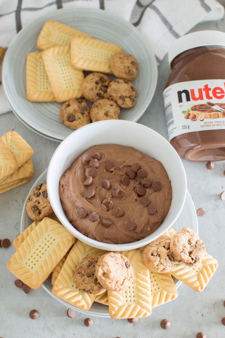 A bird's eye view of a bowl filled with Nutella cookie dip, topped off with chocolate chips, surrounded by different kinds of cookies and a jar of Nutella in the background