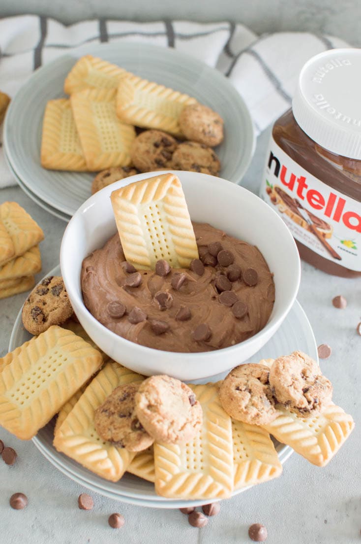 A cookie inserted into a big white bowl of Nutella Cookie Dip and chocolate chips, surrounded by cookies and a jar of Nutella in the background