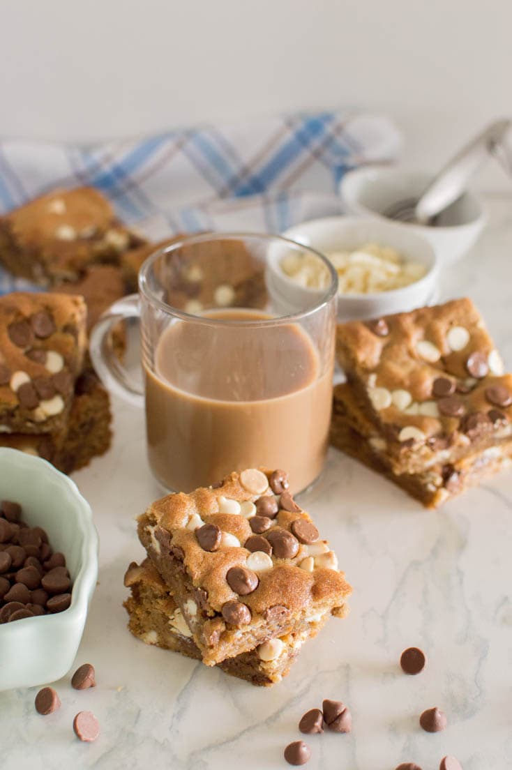 Homemade double chocolate blondies stacked on top of each other, surrounded by more slices of the blondies and with a bowl of white and milk chocolate chips and with a cup of coffee in the background
