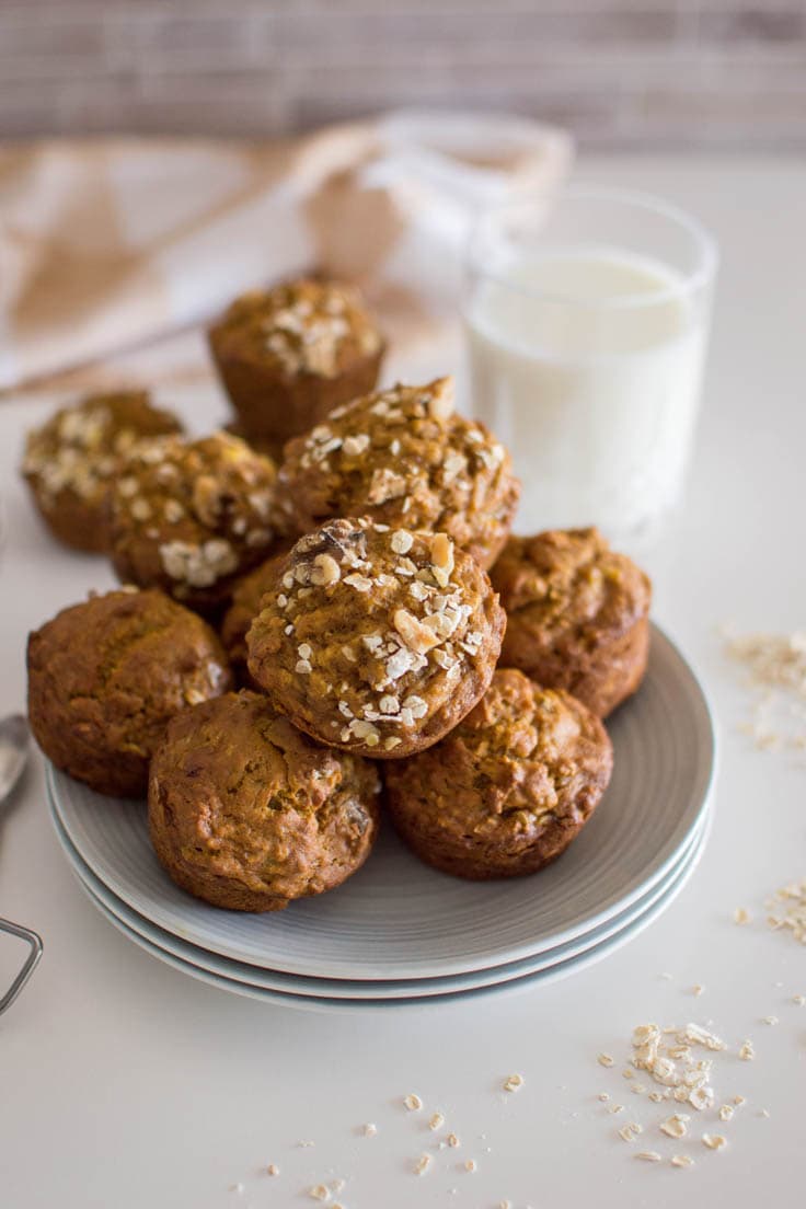 A stack of oat pumpkin muffins on a stack of grey plates. A plaid napkin and glass of milk sit in the background