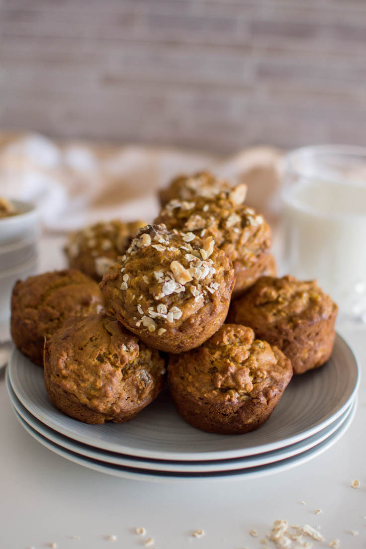 A stack of oat pumpkin muffins on a stack of grey plates. A plaid napkin and glass of milk sit in the background