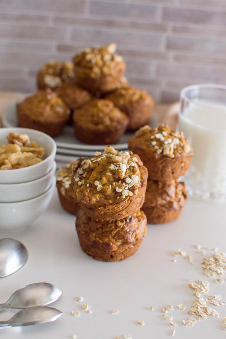 Two stacks of Oat Pumpkin Muffins with a glass of milk and a stack of grey plates with additional muffins in the background