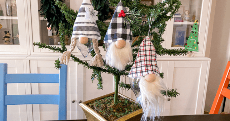 Three gnomes hanging from a small Christmas tree. The gnome of the left has a black and white striped hat with a felt flower and braids. The gnome in the middle has a black and white buffalo check hat with red berries. And the gnome on the right has a red, black and white hat.