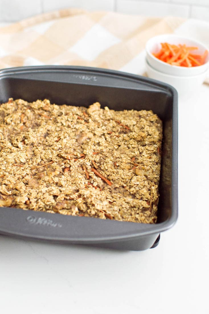 Carrot cake oatmeal bake in a dark non-stick square pan, with a bowl of shredded carrots in the background