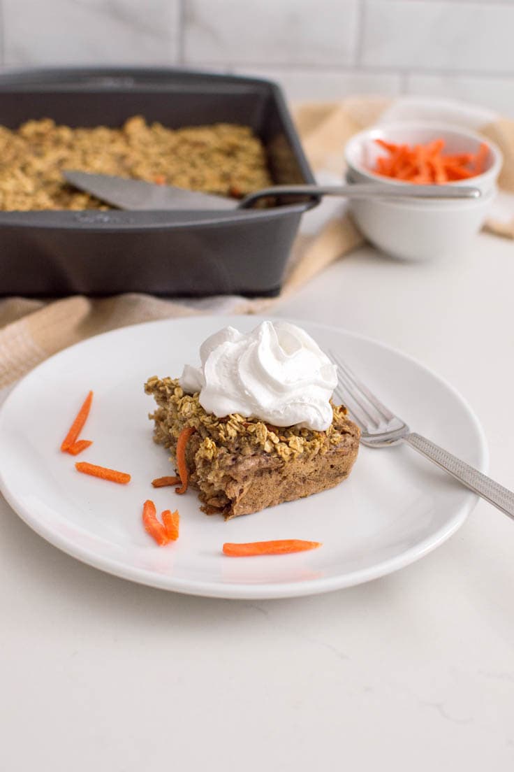 A slice of carrot oatmeal bake on a white plate, with a dollop of whipped cream and shredded carrots on the plate. A fork sits on the plate. There is a large pan of baked oatmeal in the background as well as a small bowl of shredded carrots.