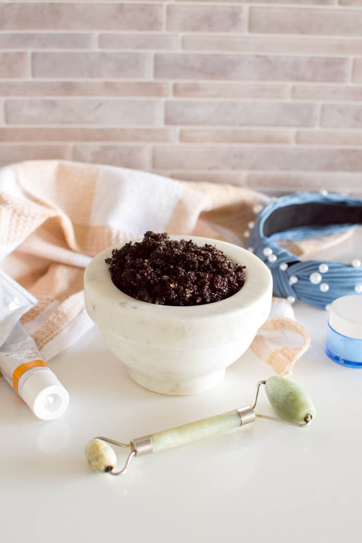 DIY Coffee Scrub in a marble bowl, surrounded by other beauty products including a jade roller, headband, and facial creams.