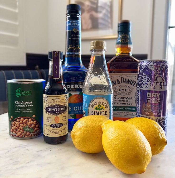 collection of the ingredients for the jack frost cocktail include garbanzo bean water, scrappy's lavender bitters, rose's simple syrup, blue curacao, Jack Daniel's Tennessee rye whiskey, lemons and dry botanical lavender bubbly.