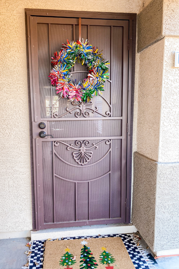 multicolored tinsel garland wreath hanging on a brown screen door with a tree door mat on the floor.