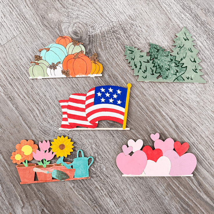 5 wood cut outs painted, top right 3 trees, top left pumpkin stack, middle American flag, bottom right hearts, bottom left flowers with watering can.
