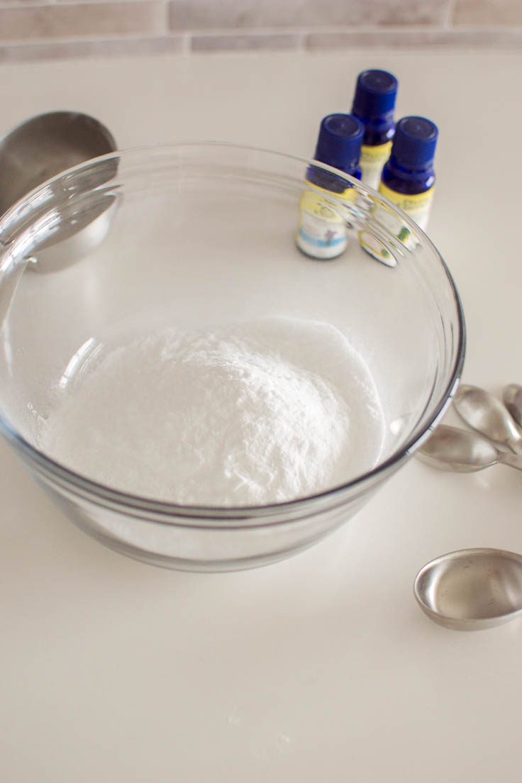 A glass bowl filled with baking soda, surrounded by measuring spoons and cups and essential oils
