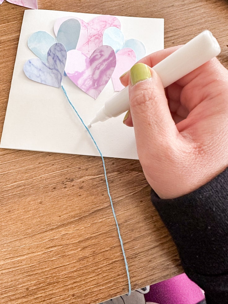 Multiple color hearts on a white card with one blue string being applied by a hand with a tube of glue