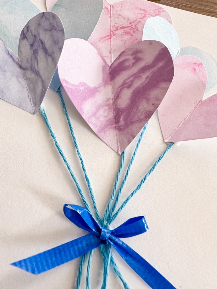 Multiple color hearts on a white card with blue strings attached to them held together by a blue bow