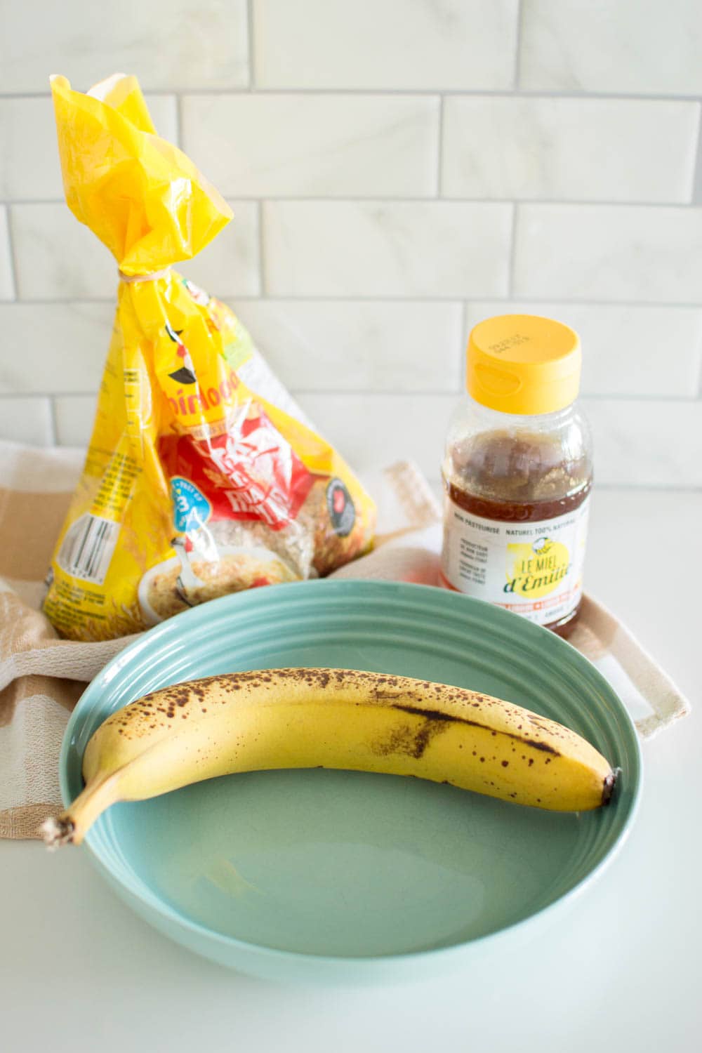 A banana sits in a green bowl, with a bag of oats and a bottle of honey in the background