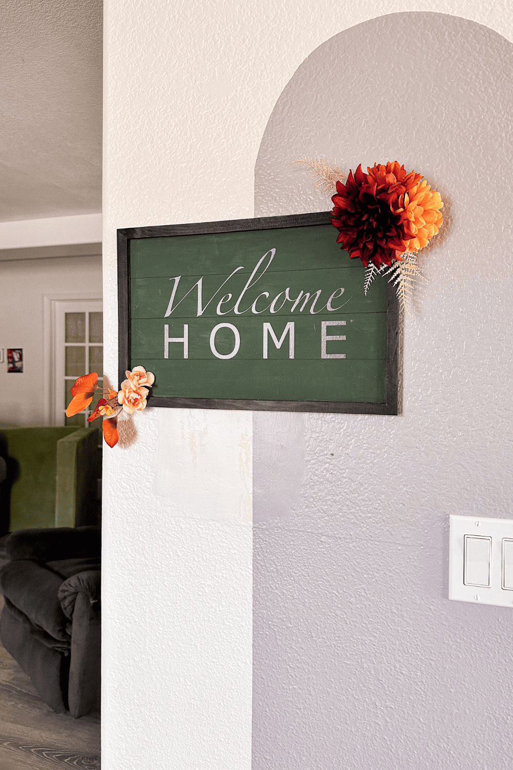 Green sign with a black frame that says Welcome Home with burgundy and burnt orange flowers hanging on a white and purple wall