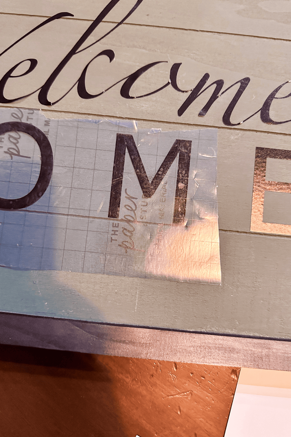 Showing a close up of transfer tape over the letter M on a green wooden sign.