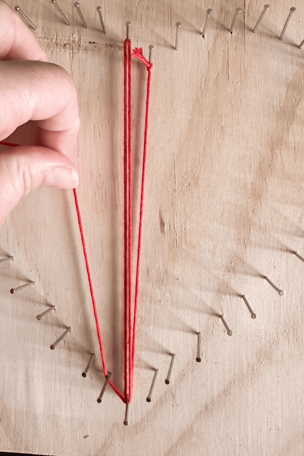 Nails on a piece of wood with string in between in red. Hand showing how to tie the strings.