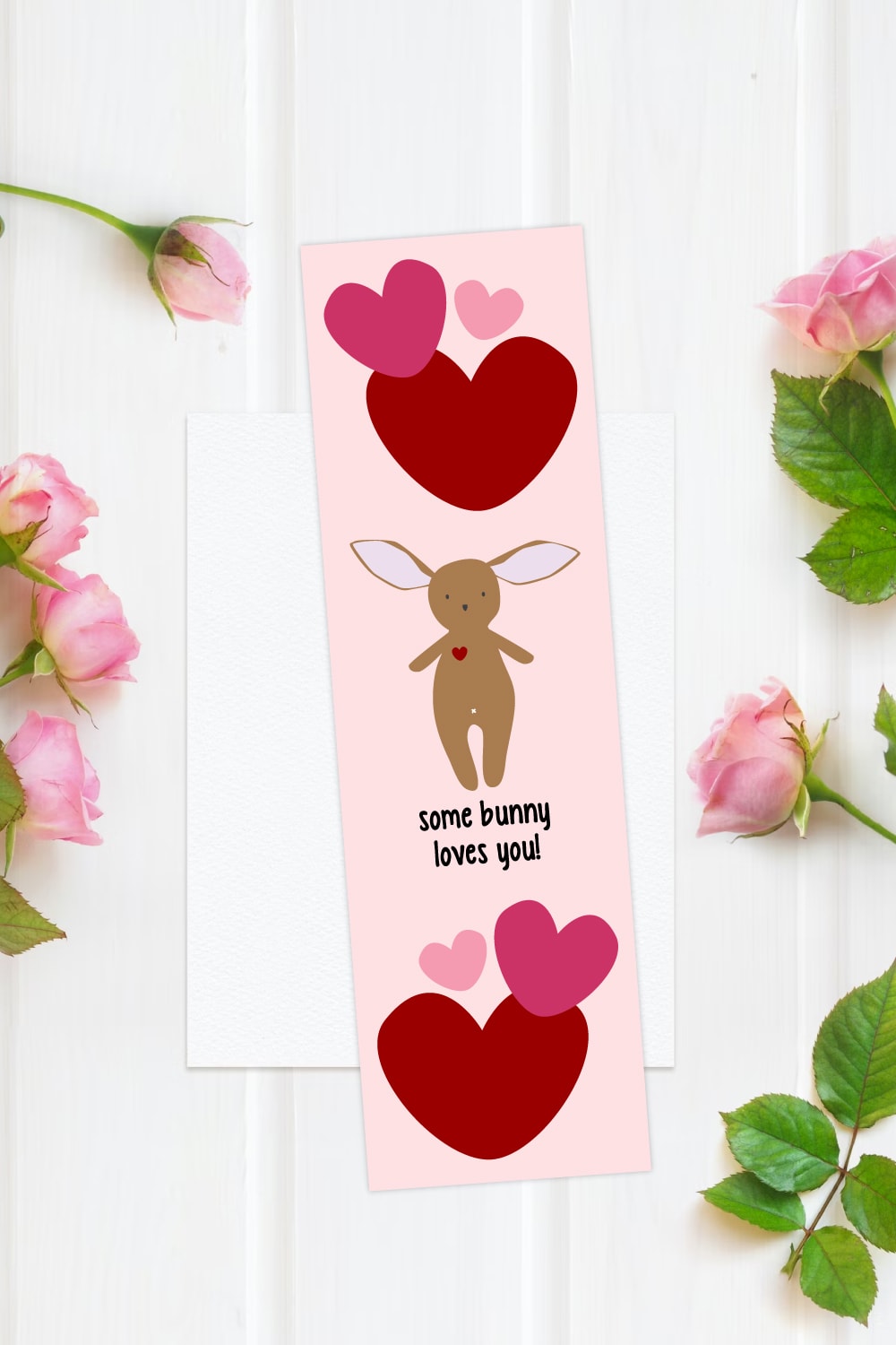 Preview of bunny valentine printable bookmark on top of white wooden background with white stationery paper and pink roses surrounding the edge border. 